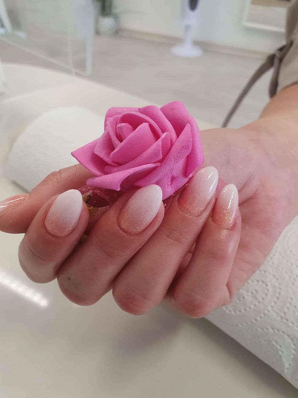 Mandelförmige Nägel mit zartem Ombré in Weiß und Rosé mit Glitzer, die Hand hält eine rosa Rose